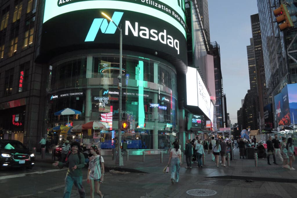 Nasdaq signage wraps the marketsite building in times square as pedestrians cross the street beneath surrounding high-rises.